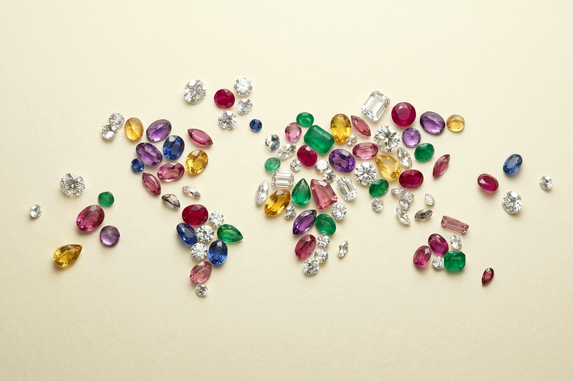 Polished diamonds and faceted multi-color gemstones (rubies, sapphires, emeralds, citrines, amethysts, pink tourmaline) scattered on warm cream paper under soft studio light