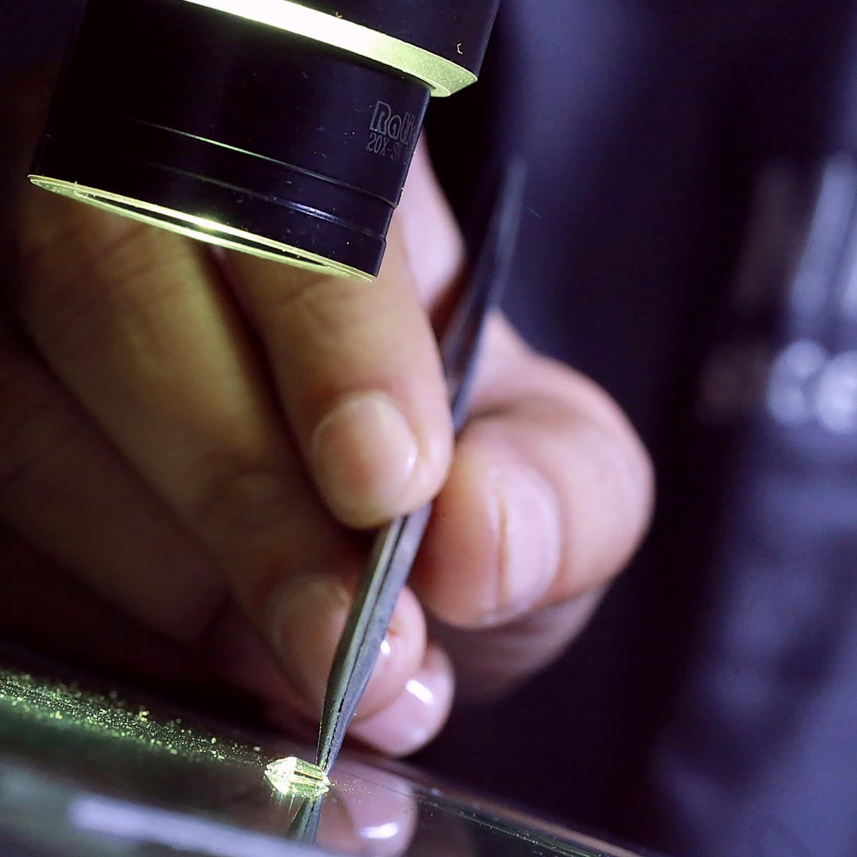 A gemmologist holding a diamond in tweezers under a loupe, wearing a NIRU coat