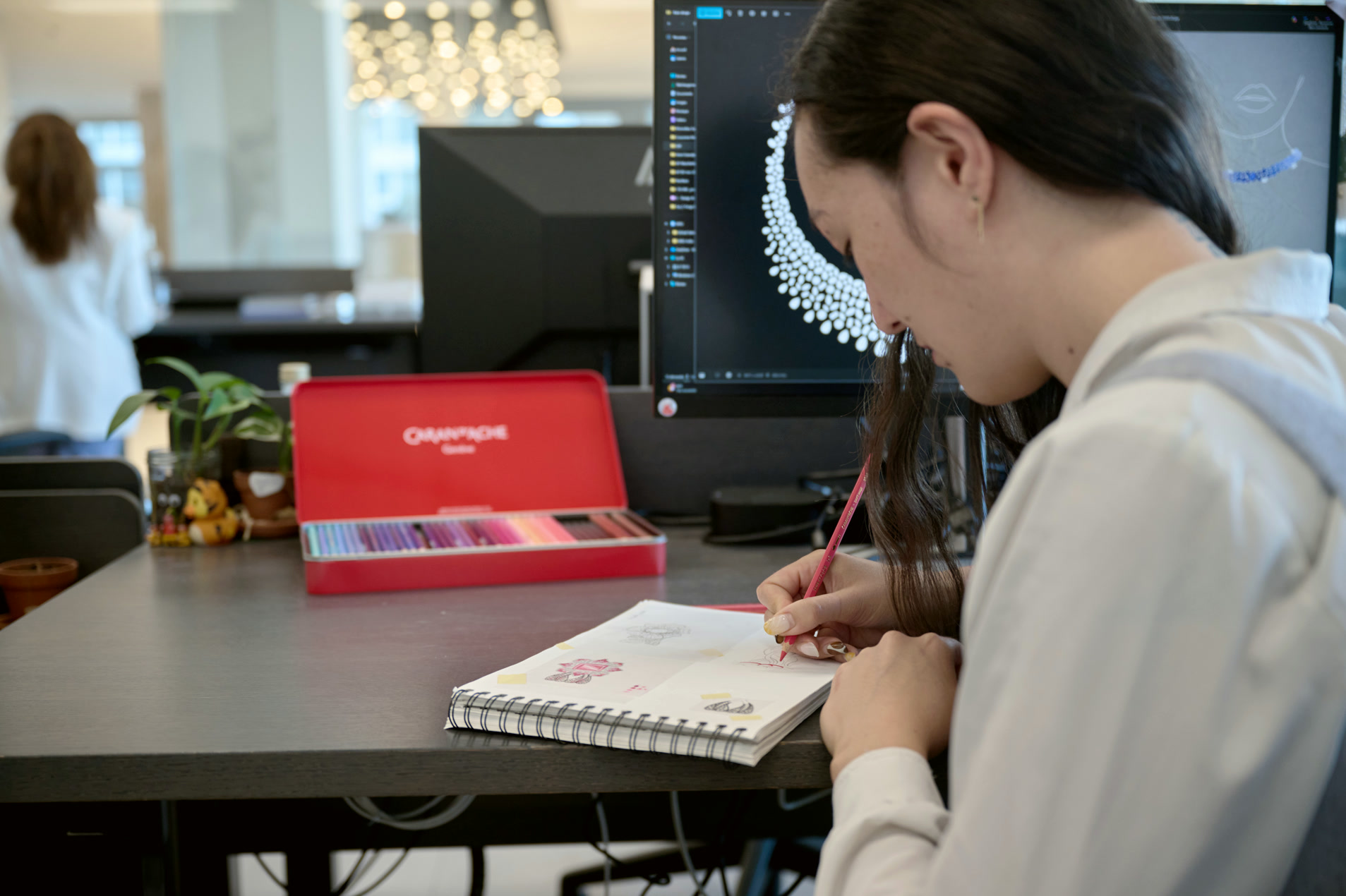 Designer sketching jewellery on paper with a finished render on the monitor behind